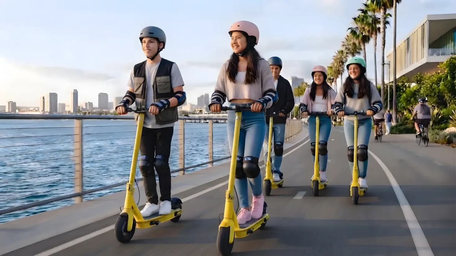 Five young people in helmets and pads ride yellow electric scooters in a line along a sunny waterfront bike path with a distant city skyline.